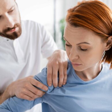 physiotherapist touching injured shoulder of redhead woman during diagnostics in consulting room